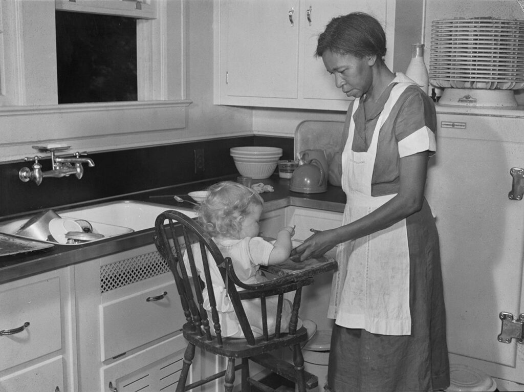 A black woman wearing a dark-colored dress and white apron feeds a white baby in a high chair in a kitchen.